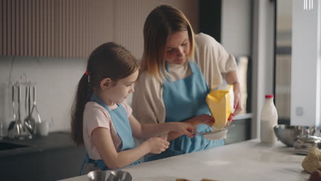 mother-is-teaching-her-little-girl-to-make-dough-and-bake-bread-in-home-woman-is-pouring-flour-in-sifter
